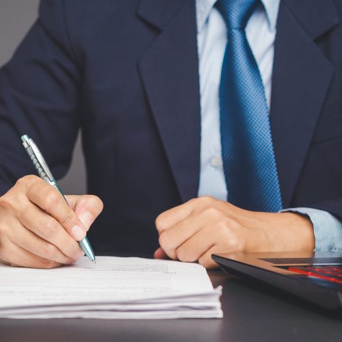 A close-up of a businessman wearing a suit and tie sitting at a desk with a laptop. He is focused on signing a document with a pen in his hand, indicating business dealings or paperwork.